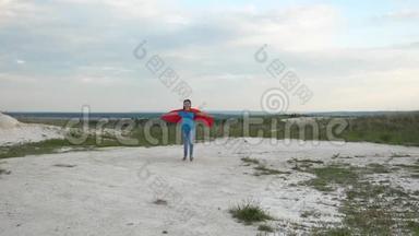 快乐女孩超级英雄穿着红色斗篷在蓝天面前跑过田野，微笑着，慢动作。 年轻女孩梦想